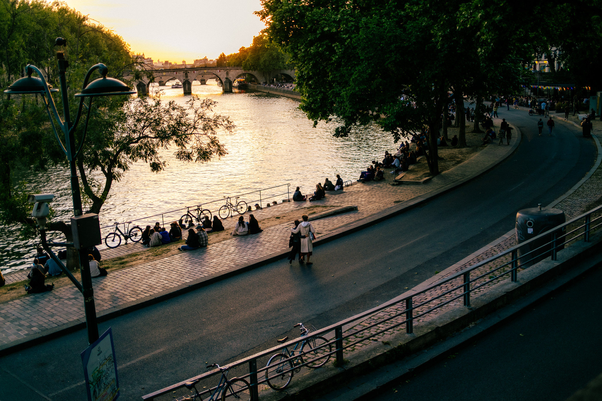 Lamp posts silhouetted above a curving quay, bicycles and figures beside gold water